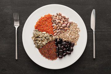 top view of fork and knife near white ceramic plate with raw assorted beans, cereals and seeds on dark wooden surface with copy space