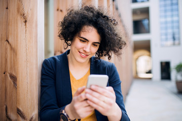 A happy young business woman with smartphone standing outdoors.