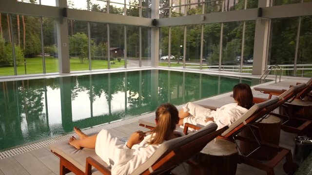 Two Woman In Bath Robes Sitting By A Large Luxurious Indoor Pool At A Cosmetic Day Spa, Chatting And Looking At The Summer Forest Through Large Windows, Back View