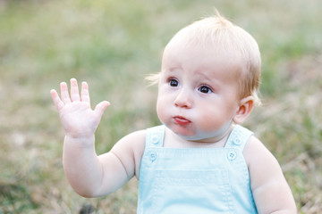 Cute blond boy sitting alone on the grass and waving his hand. The boy is ten months old