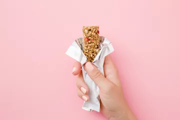 Foto op Aluminium Young woman hand holding cereal bar on pastel pink table. Opened white pack. Closeup. Sweet healthy food. © fotoduets