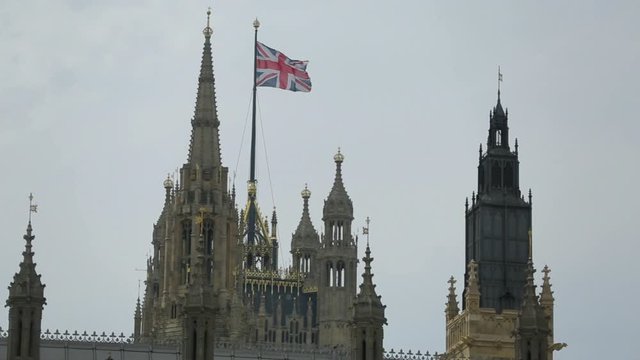 Medium low angle day time still shot of the top sharp towers of London parliament also known as Palace with British flag against clear grey sky, England, UK