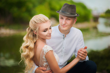 Fototapeta premium The bride with the groom in the summer on the background of textural greenery and the lake. Loving wedding couple in the city of Dnipro, Ukraine. Groom with a hat. Close-up kiss and tender hugs.