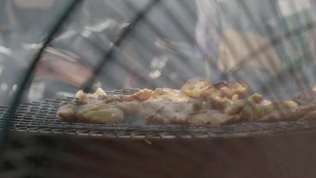 A daylight extreme closeup shot from the back of an electric fan of meat barbecues being cooked on top of a rusty makeshift charcoal grill made of iron sheet metal..