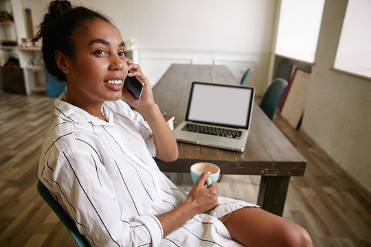 Attractive dark skinned female with bun hairstyle in striped white shirt sitting at table and giving call with her mobile phone, holding cup of coffee with charming smile