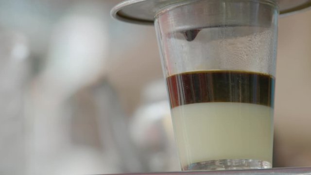 A daylight extreme closeup shot of a transparent drinking glass with milk at the bottom while collecting drip coffee from the aluminum filter on top of it..