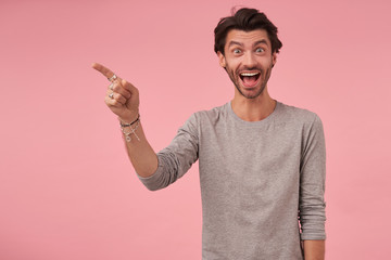 Studio shot of joyful bearded male with trendy haircut standing over pink background, wearing casual clothes, looking at camera amazedly and raising eyebrows, pointing aside with forefinger