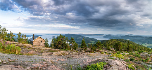 Wide panorama with public tourist rest house in front of view over wild Scandinavian mountains with pine tree forest, the village and two sea bays, summer day with heavy dramatic clouds, North Sweden