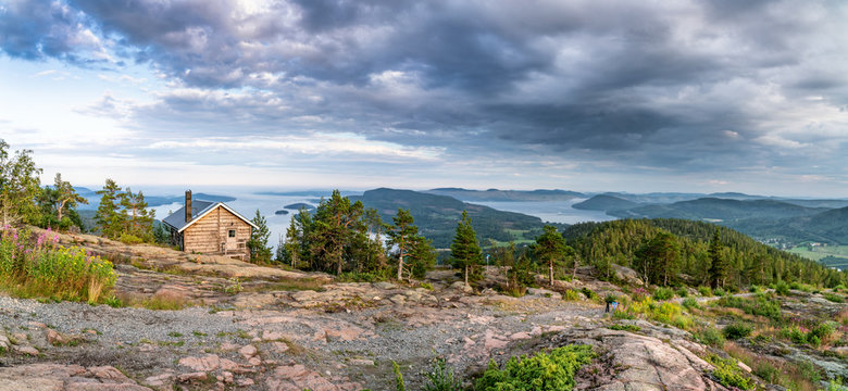 Wide panorama with public tourist rest house in front of view over wild Scandinavian mountains with pine tree forest, the village and two sea bays, summer day with heavy dramatic clouds, North Sweden