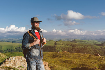 Portrait of a stylish young man with a long beard in a jeans suit, sunglasses and a cap with a backpack stands on a background of mountain valley in nature. The concept of fashion and style in nature © yanik88