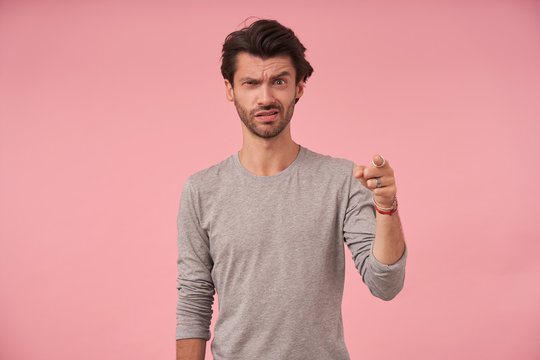 Bewildered young bearded male with trendy haircut looking at camera and frowning, standing over pink background in grey sweater, pointing ahead with index figer