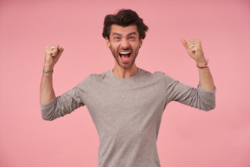 Flurried young pretty male with trendy haircut raising fists happily, standing over pink background in casual clothes, shouting joyfully and wrinkling forehead