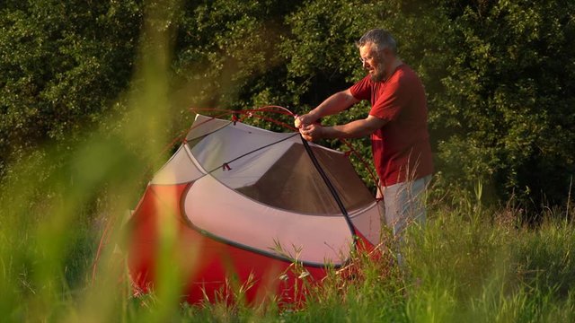 KALUGA/RUSSIA - SEPTEMBER 09 2019: Mature experienced tourist arranges small colorful tent on green grass glade against thick forest in evening on September 09 in Kaluga