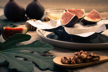 The fig leaf in the foreground and the walnut blurry. In the background is Camembert cheese with slices of figs on it. In the background are two whole figs and a transparent honey flint. Horizontal or