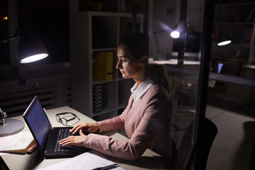 Young businesswoman siting at her workplace typing on laptop computer and working online till late at dark office