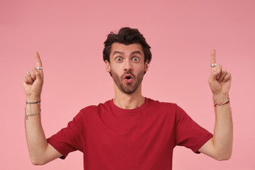 Amazed young man with beard in casual clothes standing over pink background with raised index fingers, looking at camera with wide mouth opened, contracting forehead and raising eyebrows