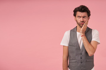 Serious young bearded man with dark hair wearing grey waistcoat and white t-shirt, standing over pink background, looking aside with pout contracting and frowning with palm on his cheek