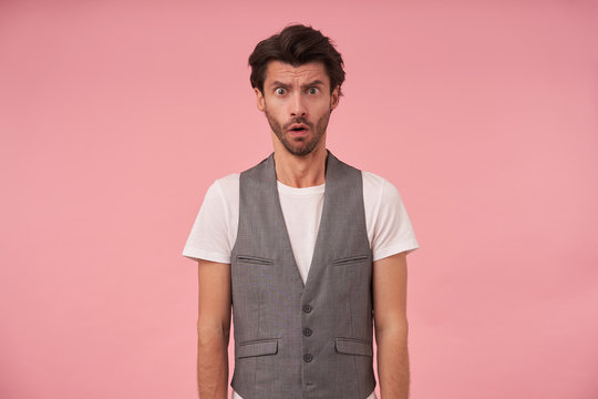 Studio photo of bearded dark male with trendy haircut in grey waistcoat and white t-shirt, posing over pink background with hands down, looking at camera with confused face, frowning with opened mouth