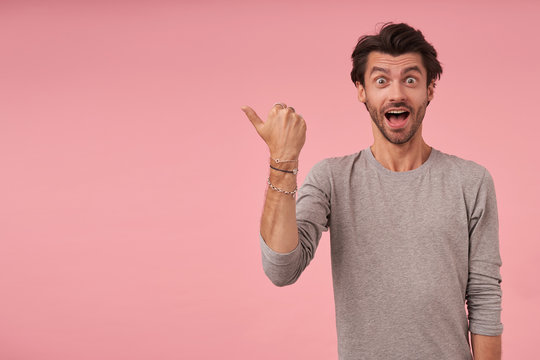 Indoor shot of excited young man with trendy haircut pointing aside with thumb, looking at camera joyfully and raising eyebrows, isolated over pink background