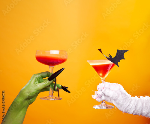 Photo of hands of mummy and zombies with wine glass and black bat on empty orange background .