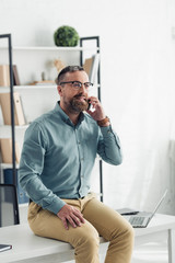 handsome businessman in shirt and glasses sitting on table and talking on smartphone