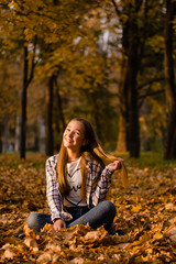 Beautiful smiling girl sitting on the ground on yellow leaves in park. Leisure time on warm autumn day
