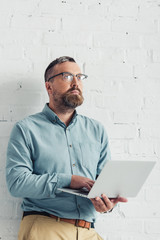 handsome businessman in shirt and glasses holding laptop and looking away