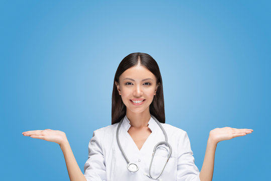 A young Asian woman doctor is scaling with her arms two different options in front of isolated and easy to remove blue background. Communicate about healthcare health and medical choices or compare 