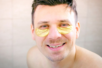 Happy young man with golden patches under eyes looking at camera and laughing, front view. He is putting himself in order making hygienic and beauty anti-aging procedures. Caring about face skin.