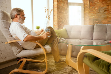 Senior man working with tablet at home - concept of home studying. Caucasian male model sitting on sofa and drinking water while serfing in internet, watching cinema or webinar, online lessons.