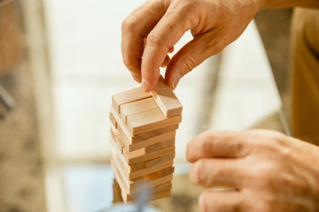 Close up of senior man's hands doing his wooden constructor at home - concept of home studying. Caucasian male model sitting on sofa and working with bricks for training of fine motor skills.