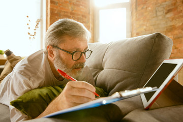 Senior man working with tablet at home - concept of home studying. Caucasian male model lying on sofa and making notes while serfing in internet, watching cinema or webinar, online lessons.
