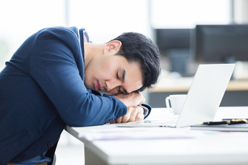 Asian young businessman worked late and fell asleep on laptop computer In the office room background.