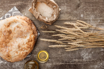 top view of lavash bread on towel with pattern near wheat spikes, rolling pin, flour and olive oil on wooden table