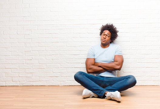 young black man shrugging, feeling confused and uncertain, doubting with arms crossed and puzzled look sitting on the floor at home