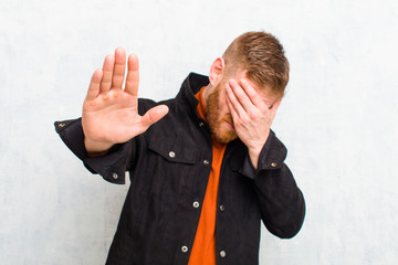 young red head man covering face with hand and putting other hand up front to stop camera, refusing...