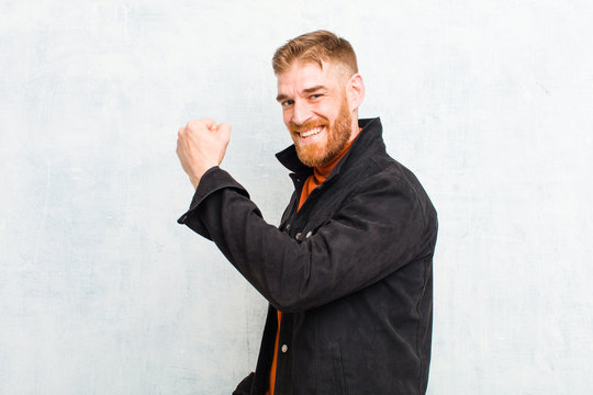 young red head man feeling happy, satisfied and powerful, flexing fit and muscular biceps, looking strong after the gym against grunge wall
