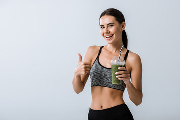 smiling sportswoman holding smoothie and showing thumb up isolated on grey