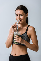 attractive smiling sportswoman drinking smoothie isolated on grey