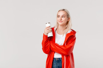 Pretty blond woman in a red raincoat holding white thermal mug with hot beverage isolated over white background.