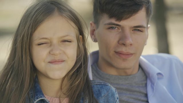 Portrait of cute little girl happily embracing with love and care her elder teenage brother in park at sunny autumn day.