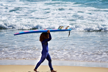 Chica paseando con la tabla de surf encima de la cabeza por una playa de arena limpia y unas olas en el mar