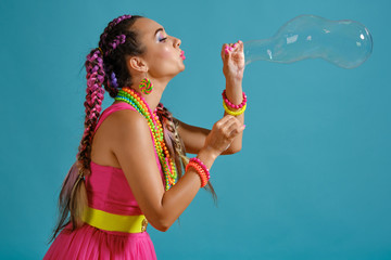 Lovely girl with a multi-colored braids hairstyle and bright make-up, is blowing bubbles using her...