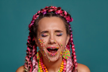 Lovely girl with a multi-colored braids hairstyle and bright make-up, posing in studio against a blue background. Colorful topping is on her face.