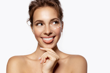 Beautiful young woman with bare shoulders looking away and applying cosmetology cream with...
