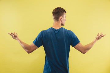 Caucasian young man's half-length portrait on yellow studio background. Beautiful male model in blue shirt. Concept of human emotions, facial expression. Showing and pointing something. Inviting.