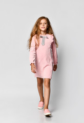 Beautiful little girl with long blond hair and blue eyes, standing on a white background, wearing a pink casual dress and gym shoes, teenager style