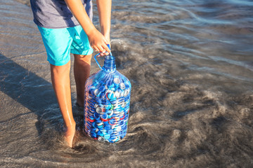 Child gathering plastic bottle caps into big water bottle at seaside. Recycling and environment friendly concept with copy space