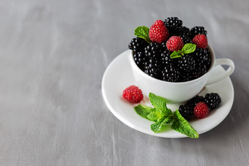 Ripe juicy raspberries, blackberries and mint in a white mug on a gray background with copy space.