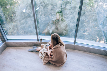Sad woman sitting on a warm floor in a socks wrapped in a woolen blanket near the large window in light. autumn mood, warmth and comfort. - Powered by Adobe